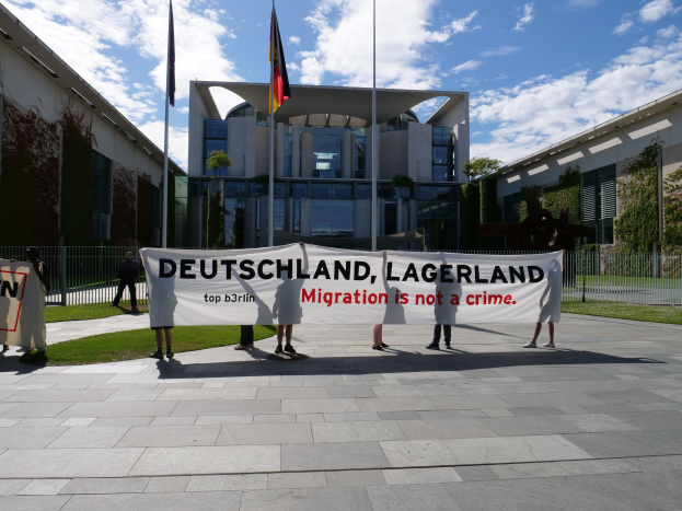 Gruppe von Menschen mit einem Transparent "Deutschland, Lagerland Migration ist kein Verbrechen" vor einem eingezäunten Gebäude mit Bäumen und Flaggen.