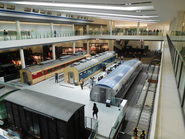 Eine große Museumsausstellung mit verschiedenen Zügen in unterschiedlichen Größen und Farben, die von Menschen auf dem Boden, Geländern und Säulen unter Deckenbeleuchtung betrachtet werden.