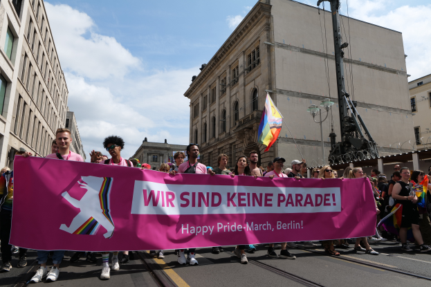 Eine Gruppe von Menschen marschiert auf einer Straße in Berlin, Deutschland, mit einer pinken "Happy Pride March"-Fahne, mit Gebäuden auf beiden Seiten der Straße und einem Fahnenmast im Vordergrund unter einem bewölkten Himmel.