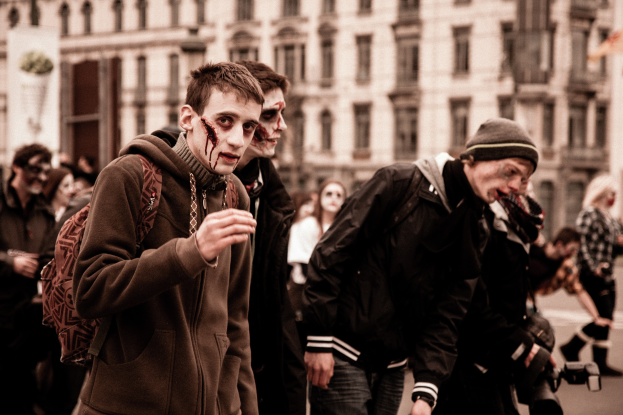 Eine Gruppe von Menschen in Zombie-Kostümen geht eine Straße entlang, wobei einige Kostüme tragen und einer eine Kamera hält, und ein Gebäude im Hintergrund.