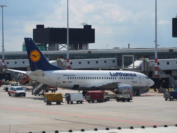 Lufthansa Airbus A320-200 am Frankfurt Airport mit Bodenfahrzeugen und Wagen im Vordergrund, einem Geb"ude und Laternen im Hintergrund, unter einem klaren blauen Himmel.