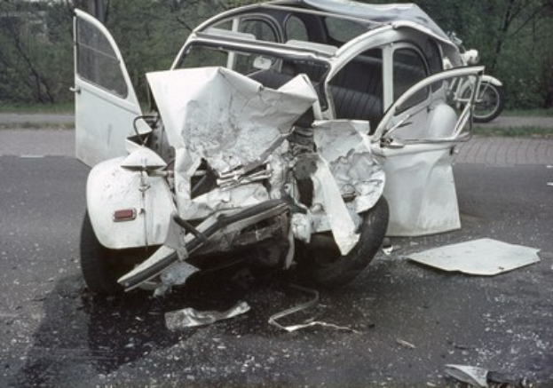 Auto mit schwer beschädigtem Frontteil am Straßenrand, umgeben von Bäumen und einem Gebäude, mit einem weiteren Fahrzeug im Hintergrund.