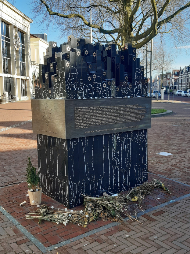 Denkmal in einem Stadtplatz gewidmet an Holocaust-Opfer mit einem Baum davor, einem Topfgewächs auf dem Boden, verstreuten trockenen Blättern und Gebäuden, Fahrzeugen, Pfählen und Bäumen im Hintergrund.