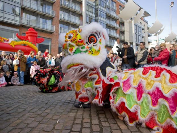 Ein lebendiges chinesisches Neujahrsfest in Amsterdam mit einer Löwen-Tanz-Show vor einer Menschenmenge, darunter einige mit Kameras, vor einer Kulisse aus Gebäuden, Laternenmasten und einem klaren blauen Himmel.