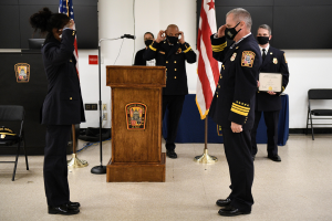 Gruppe uniformierter Personen in Masken vor einem Podium während einer Amtseinführung für einen neuen Feuerchef, der ein Zertifikat hält, mit Flaggen und Sitzplätzen.
