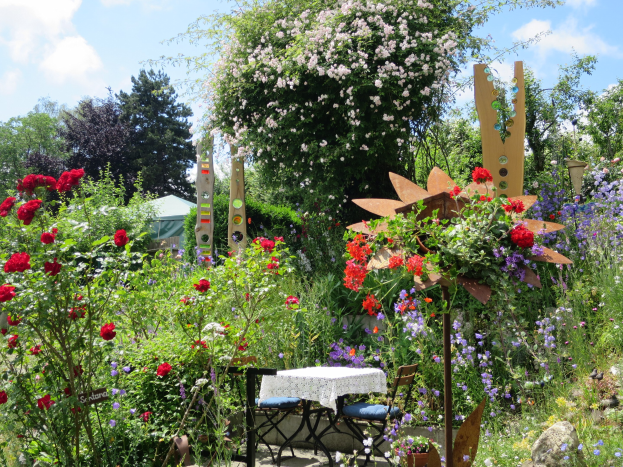 Ein Garten mit bunten Blumen und Pflanzen, der einen zentralen Tisch mit einer Decke und Stühlen zeigt, vor einem Baum und einem bewölkten Himmel auf der Chelsea Flower Show.