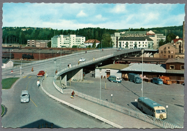 Altes Schwarz-Weiß-Foto einer Stadtstraße mit Autos, Bussen, Fußgängern auf einer Brücke, Laternenpfählen, mehrstöckigen Gebäuden mit Fenstern, Bäumen und einem bewölkten Himmel.