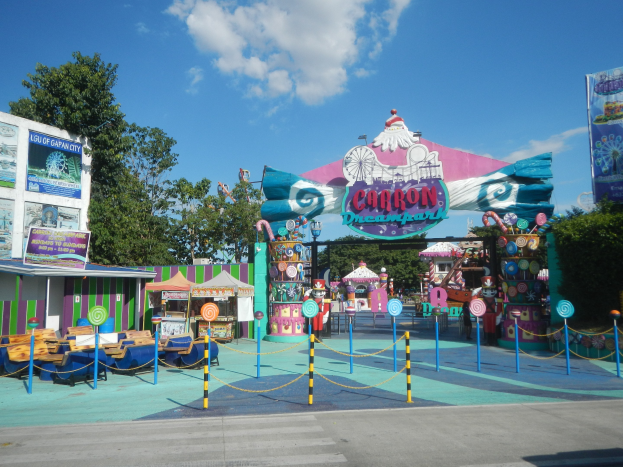 Kirmesfahrzeug am Eingang eines Parks mit einer Straße, einem Zaun, Buden, Schildern, Bäumen und einem bewölkten Himmel im Hintergrund.