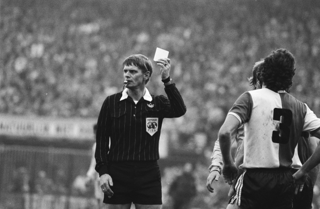 Schwarzes Bild eines Schiedsrichters mit roter Karte vor einer Menge in Fußballuniformen mit einem Zaun im Hintergrund.