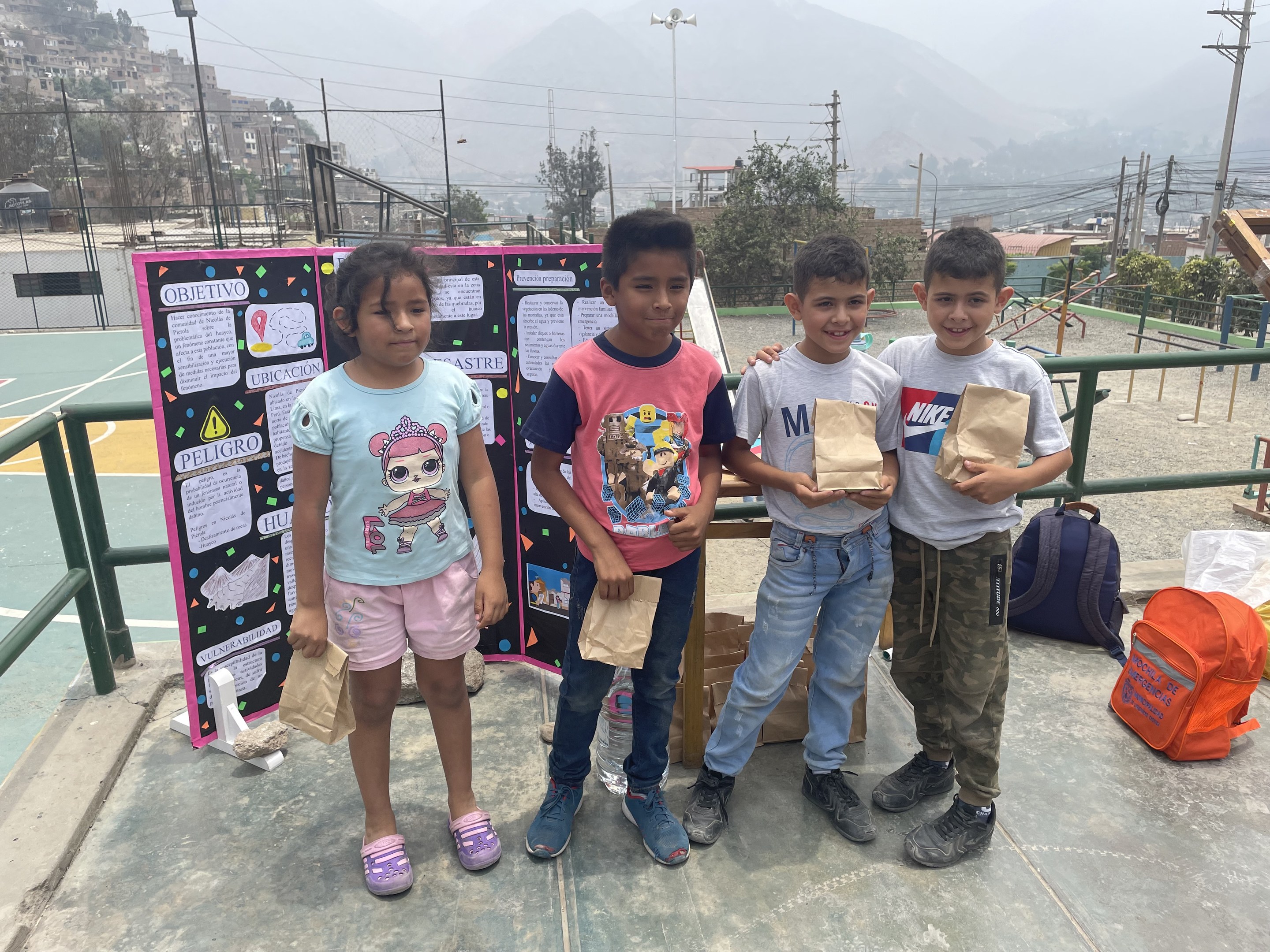 Gruppe von Kindern auf einem Basketballfeld, die Papiertüten halten, mit Tüten und Gegenständen rechts und einer Tafel mit Text links, vor einem Hintergrund aus Zäunen, Pfählen, Straßenlaternen, Bäumen, Gebäuden, Hügeln und Himmel.