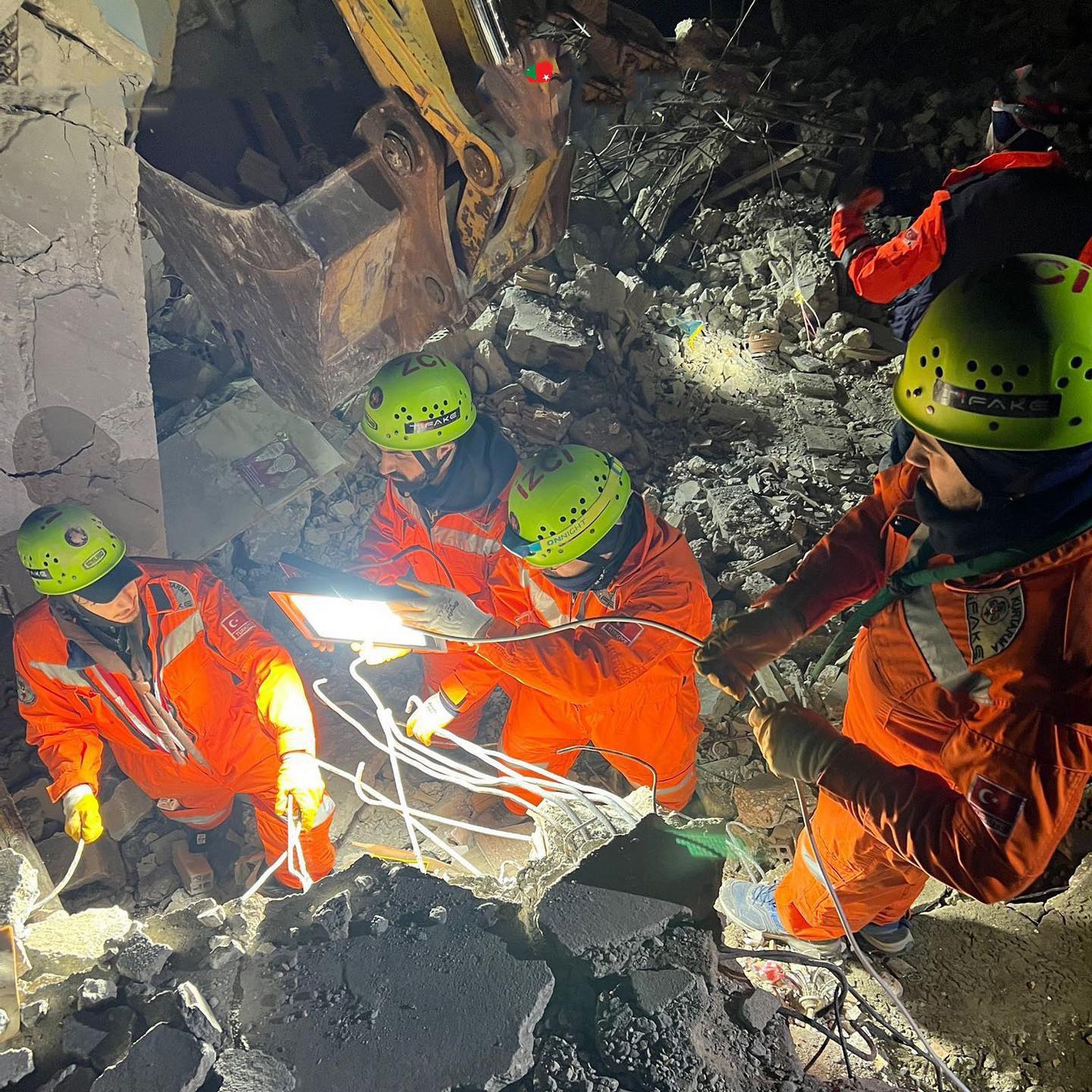 Eine Gruppe von Menschen in orangefarbigen Uniformen und grünen Helmen bei der Arbeit an Schutt mit einem Kran im Hintergrund, die Rettungsgeräte und Materialien halten.