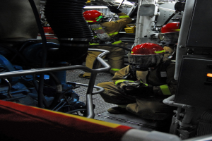 Feuerwehrleute in Helmen und Schutzausrüstung arbeiten an einem Löschfahrzeug mit Schläuchen und einer Leuchte im Hintergrund.
