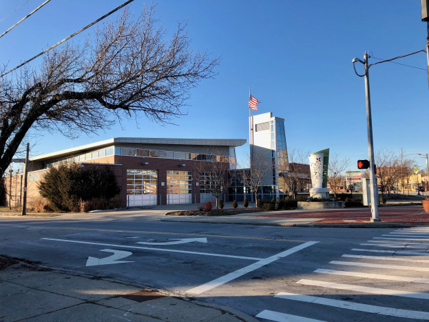 Großes Gebäude mit einer Flagge auf dem Dach, identifiziert als neue Feuerwache an einer Straßenkreuzung, umgeben von Bäumen, Straßenmöblierung und sichtbarem Himmel.