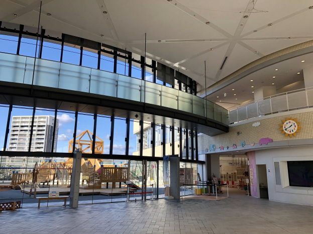 Innenansicht eines großeen Gebäudes mit zahlreichen Fenstern, Deckenbeleuchtung, einer Wanduhr, einem Fernseher, einem Anzeigebildschirm, hölzernen Konstruktionen, einem hölzernen Zaun, benachbarten Gebäuden, Bäumen und einem bewölktem Himmel, das als Haupteingang zu einem neuen Kinderspielplatz dient.