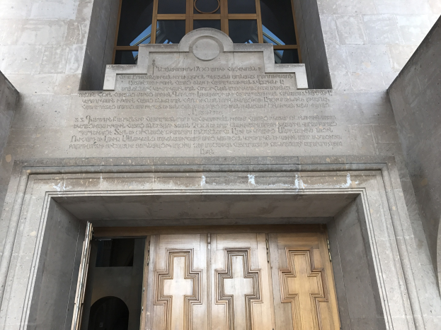 Große hölzerne Tür vor einem steinernen Gebäude mit einem Glasfenster oben, mit eingravierter Inschrift auf der Wand, die wahrscheinlich Holocaust-Opfer gedenkt.