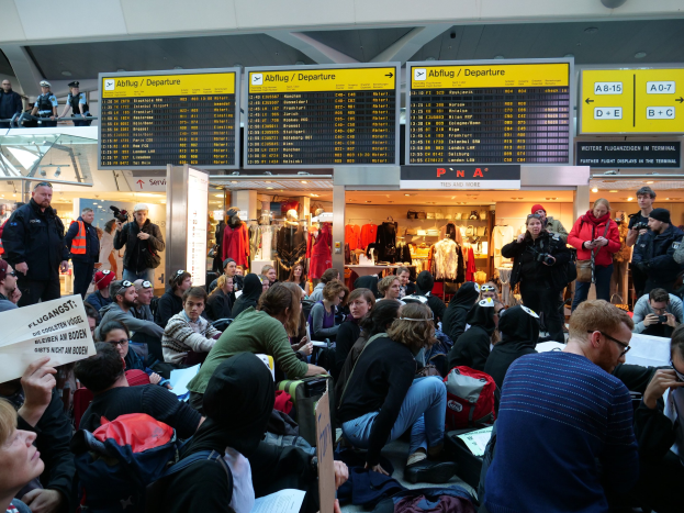 Eine große Gruppe von Menschen, die auf einem Flughafen während einer Demonstration sitzen und stehen, mit Informationsschildern, Schaufensterpuppen in Kleidern und Deckenbeleuchtung im Hintergrund.