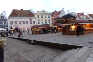 Ein lebendiger Weihnachtsmarkt in Tallinn, Estland, mit Menschen um beleuchtete und dekorierte Stände, Gebäuden mit Fenstern im Hintergrund, Treppen, die zum Markt führen, und Töpfen mit Pflanzen drumherum.