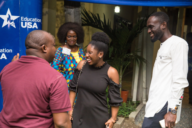 Gruppe von Menschen, die vor einem blauen Zelt mit einem Banner, Topfpflanzen, einer Säule, einer Wand und einer Decke mit Leuchten sprechen.