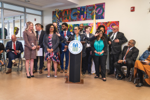 Gruppe von Menschen vor einem Rednerpult mit Mikrofon und einer Tafel mit der Aufschrift "Sancho-Stadtrat tritt dem Stadtrat bei", mit sitzenden und stehenden Teilnehmern, Fahnen, Feuerlöscher und Deckenbeleuchtung im Hintergrund.