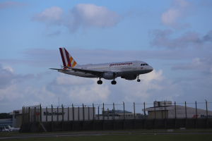 Germanwings Airbus A320-200 startet von Frankfurt Airport mit Gras darunter, einem Zaun im Vordergrund, Gebäuden und Bäumen im Hintergrund und einer bewölkten Himmel darüber.