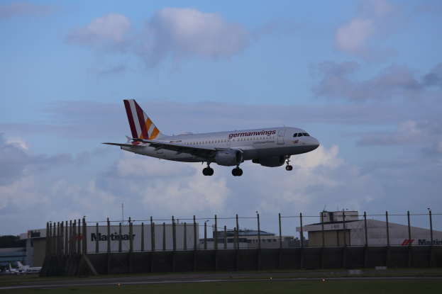 Germanwings Airbus A320-200 startet von Frankfurt Airport mit Gras darunter, einem Zaun im Vordergrund, Gebäuden und Bäumen im Hintergrund und einer bewölkten Himmel darüber.