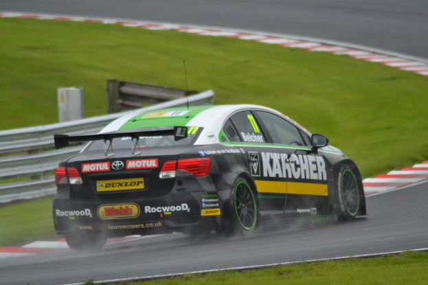 Ein Auto mit Text auf der Seite fährt auf einer regennassen Rennstrecke, die von Gras gesäumt ist, mit einer Reling und einem hölzernen Gebilde im Hintergrund.