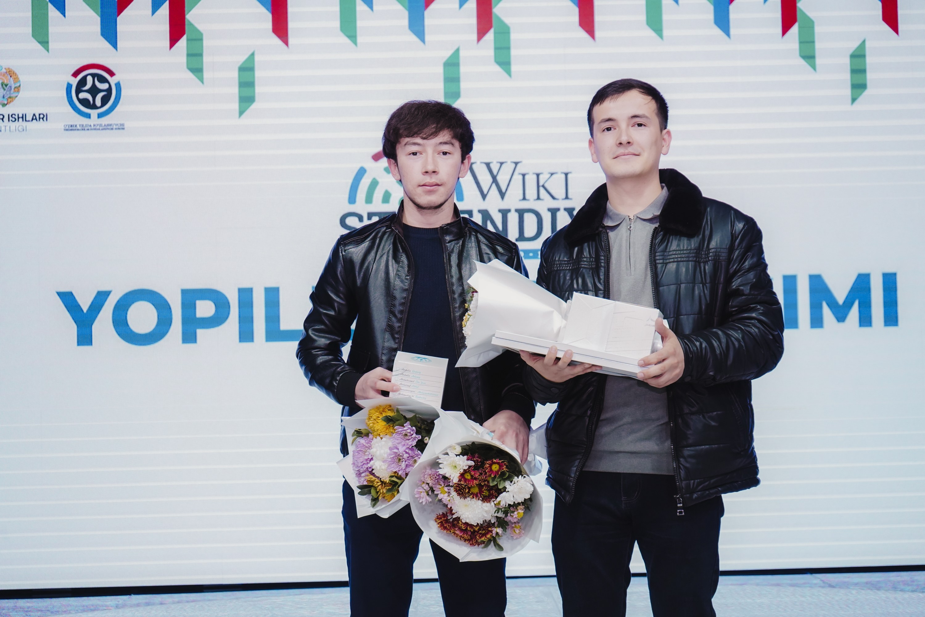Zwei Männer in schwarzen Jacken mit Blumensträußen stehen auf einer Bühne neben einem Banner mit der Aufschrift 'Yopil International Film Festival.'