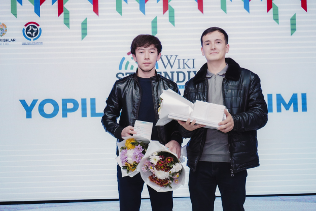 Zwei Männer in schwarzen Jacken mit Blumensträußen stehen auf einer Bühne neben einem Banner mit der Aufschrift 'Yopil International Film Festival.'