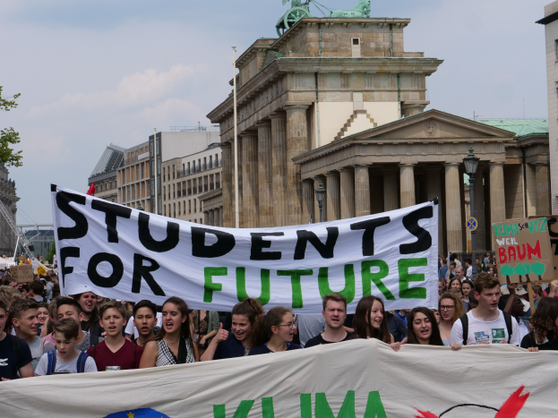 Eine Gruppe von Schülern marschiert in Berlin, hält eine bunt bemalte 'Students for Future'-Schleife gegen eine Kulisse aus Gebäuden, Bäumen und Himmel.