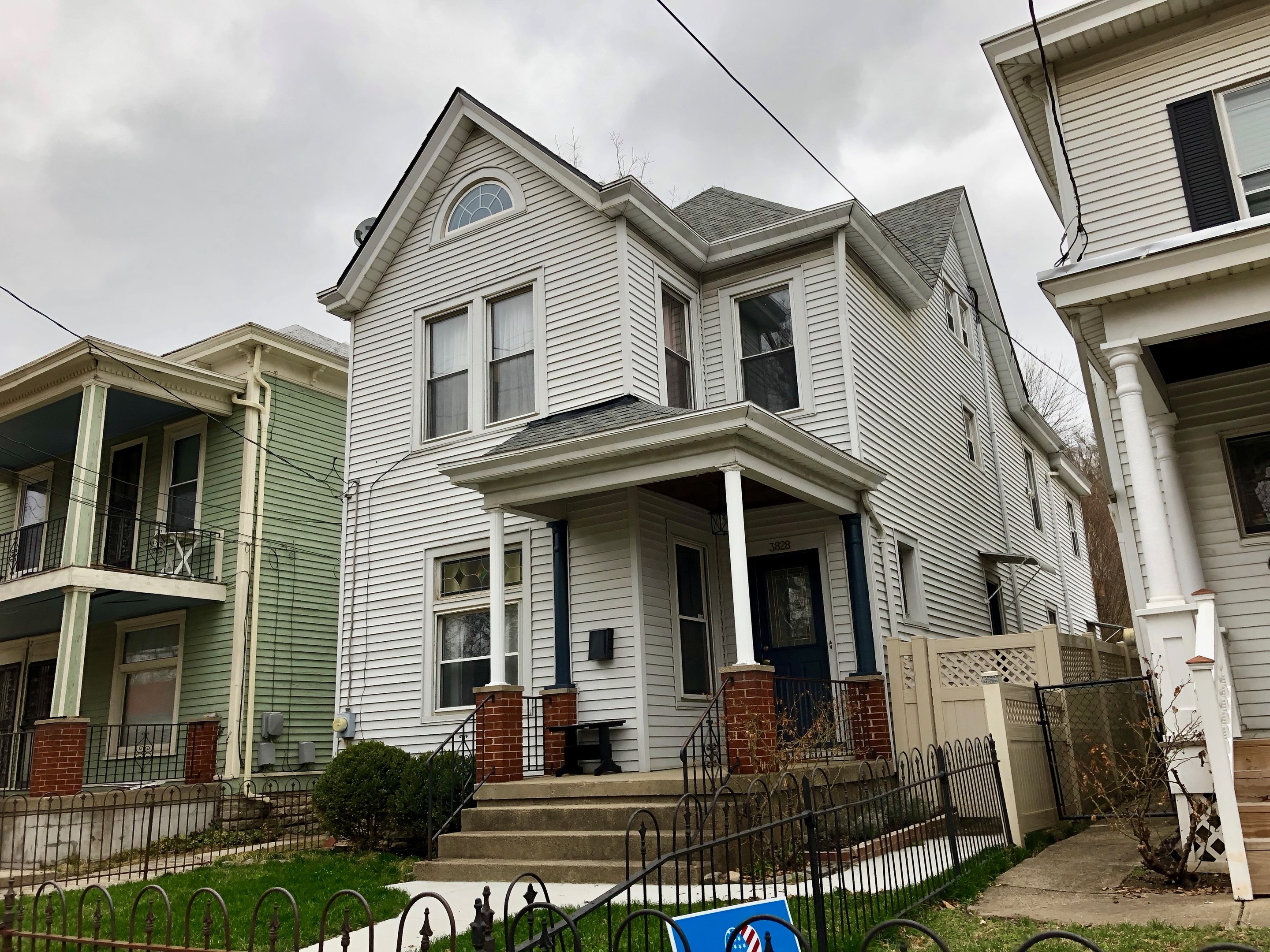 Reihe von Häusern mit Fenstern, Türen, Säulen, Geländern, Stufen und Dächern, umgeben von einem Metallzaun, Gras, Pflanzen, Bäumen und einem bewölkten Himmel, mit einem Mietschild am Boden, das die Adresse als 5901 S Elmwood Ave, Chicago 60618 angibt.