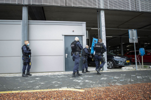 Gruppe von Polizeibeamten vor einem Gebäude mit einer Tür in einem Parkplatz stehend, umgeben von Fahrzeugen, Pfählen, Brettern und verstreuten Steinen.