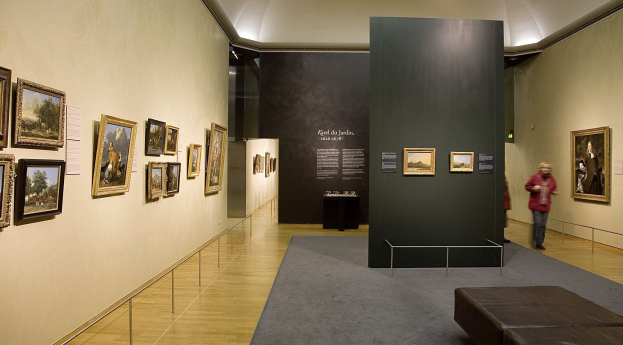 Ein großer Raum mit zahlreichen paintings an den Wänden, mit zwei Personen, einer Couch, einem Tisch mit Gegenständen und einer Tafel, die "Karel du Jardin Ausstellung in der Nationalgalerie" trägt, unter Deckenbeleuchtung.