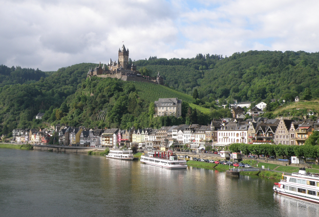 Ein malerischer Blick auf den Rhein in Deutschland, mit einer Burg auf einem Hügel, Booten auf dem Fluss, Fahrzeugen auf einer nähergelegenen Straße und einem bewölktem Himmel.