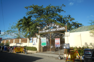 Außenansicht eines Schulgebäudes mit einem "Willkommen"-Schild, Fahrzeugen auf der Straße, bepflanzten Blumen, Informationsschildern, einem verschlossenen Namensschild, Bäumen, Gebäuden und einem bewölkten Himmel.