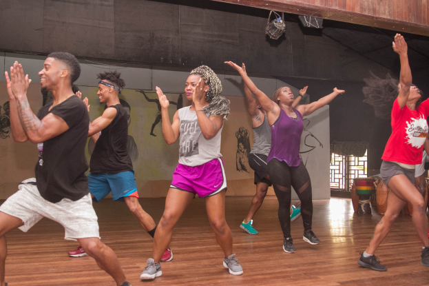 Gruppe von Menschen, die in einem Studio mit einer bemalten Wand, Fenster und Deckenbeleuchtung tanzen, wahrscheinlich während eines Zumba-Kurses.