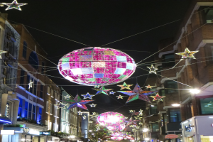 Eine festlich geschmückte Stadtstraße bei Nacht mit zahlreichen Lichtern und Dekorationen, darunter ein großer Diskokugel, der von einem Gebäude hängt.