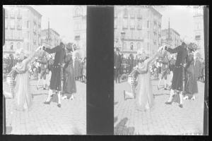 Schwarzes und weißes Foto von Menschen in Kostümen, die auf einer Straßenparade tanzen, umgeben von Gebäuden, Straßenmasten und Lichtern unter einem klaren Himmel.