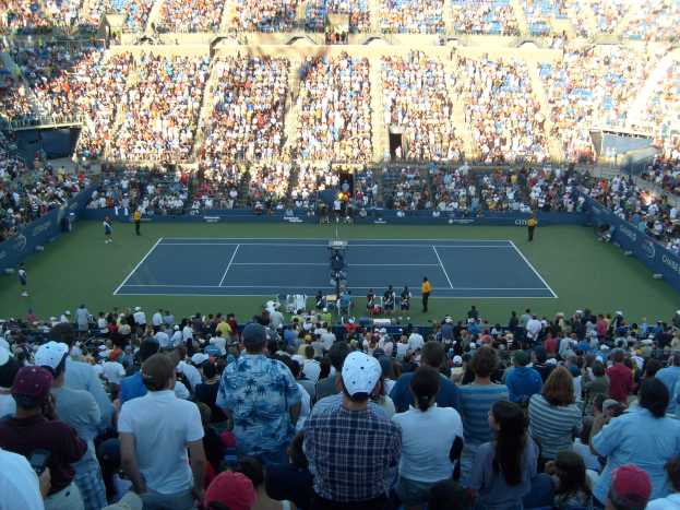 Große Zuschauermenge bei einem Tennisspiel in einem ausverkauften Stadion mit Spielern auf dem Platz und Zuschauern in den Rängen.