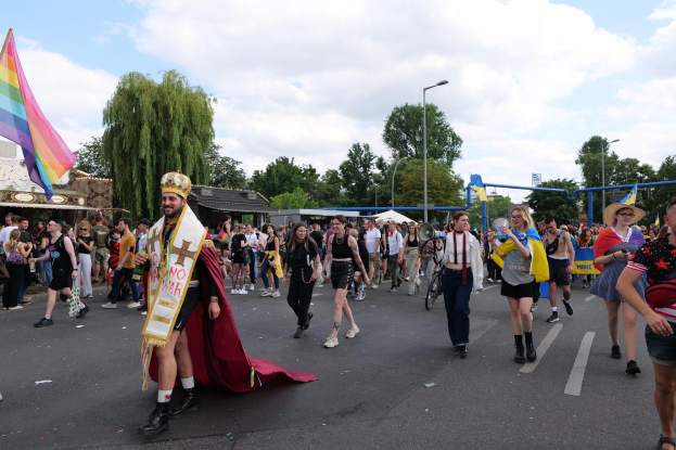 Eine Gruppe von Menschen bei der 2018 Gay Pride Parade mit einer Regenbogenflagge und Musikinstrumenten, einige tragen Mützen, vor einem Hintergrund von Laternenmasten, Bäumen, Schuppen und einem bewölkten Himmel.