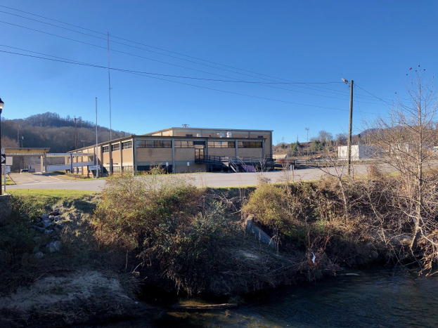 Ein Fluss durchquert eine grüne Wiese neben einem Gebäude in der Nähe von Bäumen, Pflanzen, Gras, Steinen, Straßenlaternen, Strommasten, Kabeln, Hügeln und einem klaren blauen Himmel, wobei das Gebäude für eine Wasseraufbereitungsanlage geplant ist.