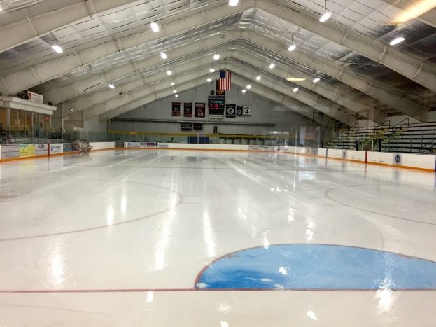 Große Indoor-Eisfläche mit Deckenbeleuchtung, Werbetafeln, einer Wand, Treppen mit Geländern und einigen Menschen, was auf eine Sportanlage hinweist, die likely für Hockey genutzt wird.