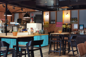 Innenansicht eines Restaurants mit Holztischen, Stühlen, einer Topfpflanze auf einem Tisch, einer Schiefertafel, einem Kühlschrank, einem Feuerlöscher, Regalen mit Flaschen, Schränken mit Gegenständen, einem gerahmten Foto an der Wand, Deckenleuchten und Lampen.