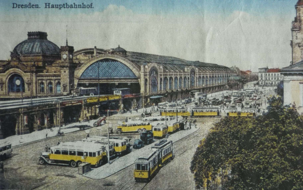 Schwarz-weiß-Postkarte des Dresdner Hauptbahnhofes: Ein belebtes Straßenbild mit Fahrzeugen, Fußgängern, Bäumen, Gebäuden und einem bewölkten Himmel.