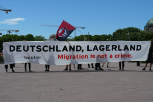Eine Gruppe von Menschen, die draußen stehen und eine Fahne halten, auf der steht "Deutschland, Lagerland Migration ist kein Verbrechen" und eine Fahne, mit Straßenlaternen, Bäumen, Kränen, einem Gebäude und einem bewölkten Himmel im Hintergrund.