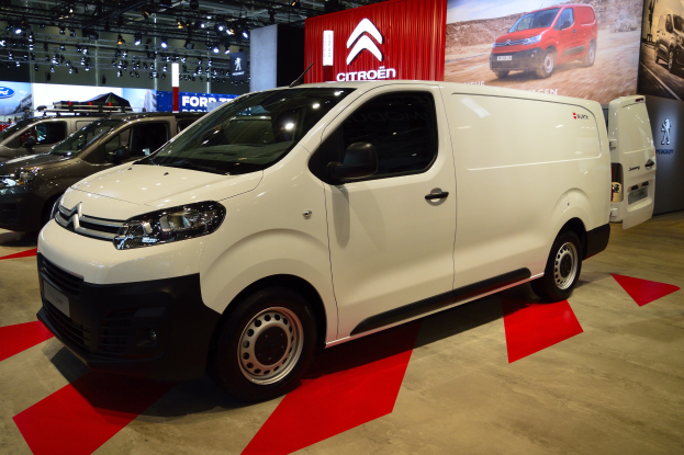 Ein weißer Citroën Berlingo Van wird auf einer Automesse präsentiert, umgeben von schwarzer Schlafzimmer-Möbeln, mit Wänden, die mit Brettern und Lichtern geschmückt sind.