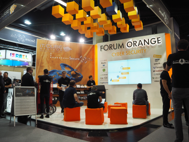 Eine Gruppe von Menschen an einem IT-Cube Systems Forum Orange Cyber Security Stand, mit sitzenden und stehenden Teilnehmern, einem Anzeigebildschirm, Texttafeln, Regalen mit Gegenständen, Deckenbeleuchtung und Lampen.