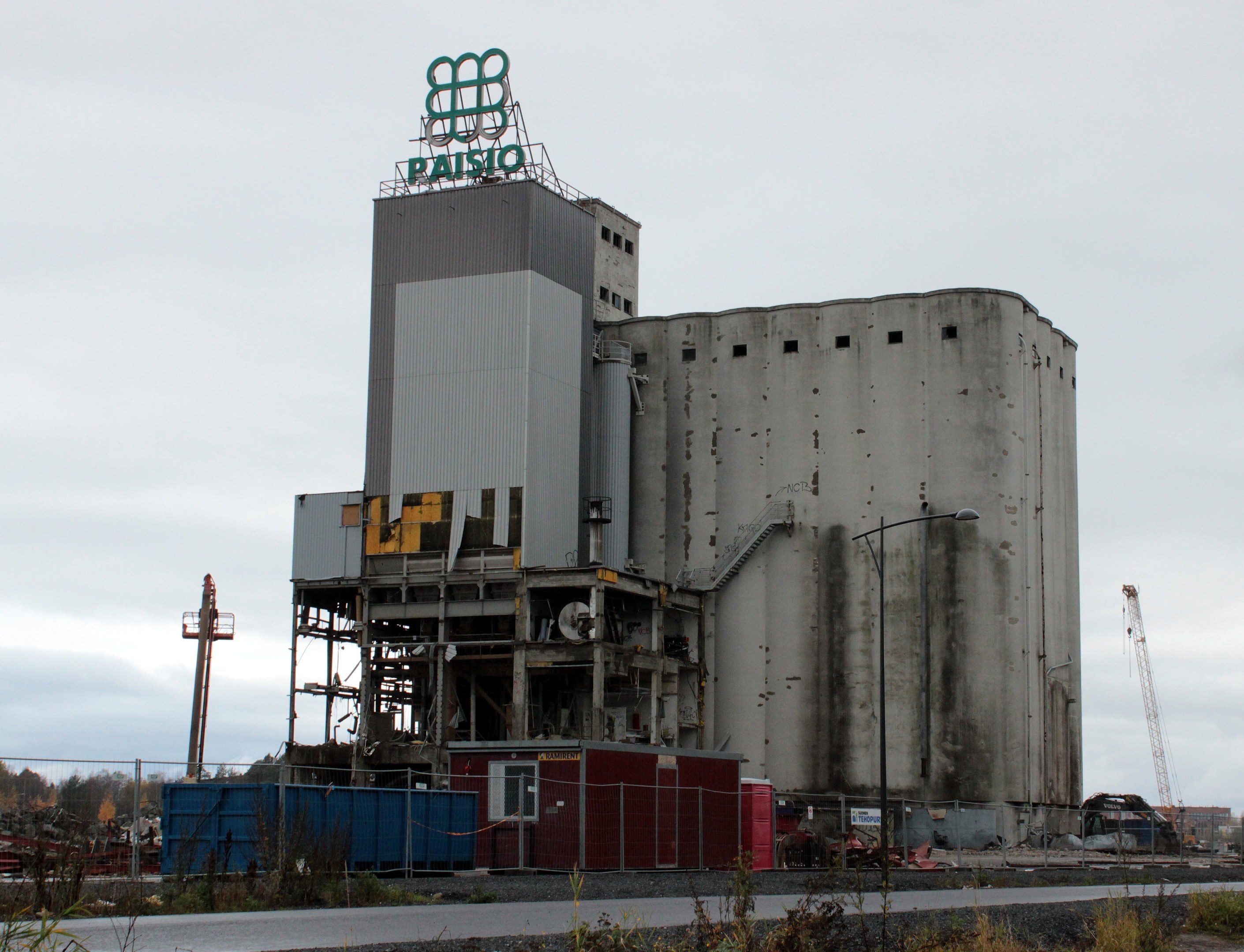 Großes Zementgebäude neben einer Straße, umgeben von Vegetation, einem Zaun, Strommasten und Lampen, mit Bäumen und einem klaren blauen Himmel im Hintergrund.