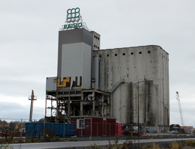 Großes Zementgebäude neben einer Straße, umgeben von Vegetation, einem Zaun, Strommasten und Lampen, mit Bäumen und einem klaren blauen Himmel im Hintergrund.