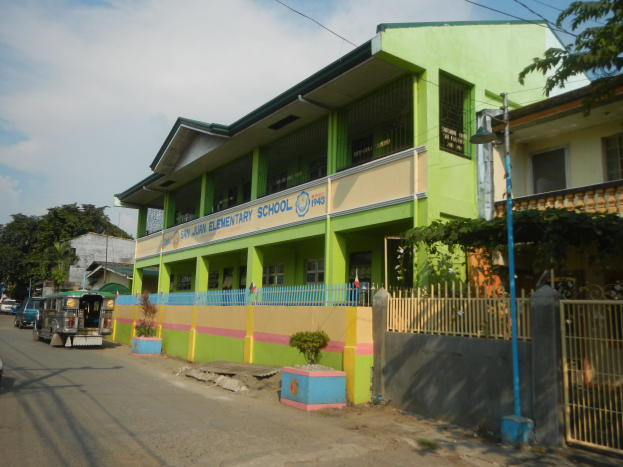 Schule mit grüner und gelber Fassade, Straße mit Fahrzeugen, Topfpflanzen, Zaun, Tor, Straßenlaterne, Bäume, Drähte und bewölkter Himmel.