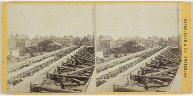 Schwarz-weiß-Foto einer Zugschiene mit Gebäuden im Hintergrund, Text 'Merkelbach & Co, Co. Amsterdam' rechts und verstreute hölzerne Gegenstände vorn.